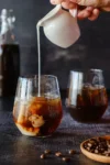 Cold Brew Kaffee: zwei Gläser mit Cold Brew und Eiswürfeln, daneben Kaffeebohnen. In ein Glas wird gerade Milch eingeschenkt.